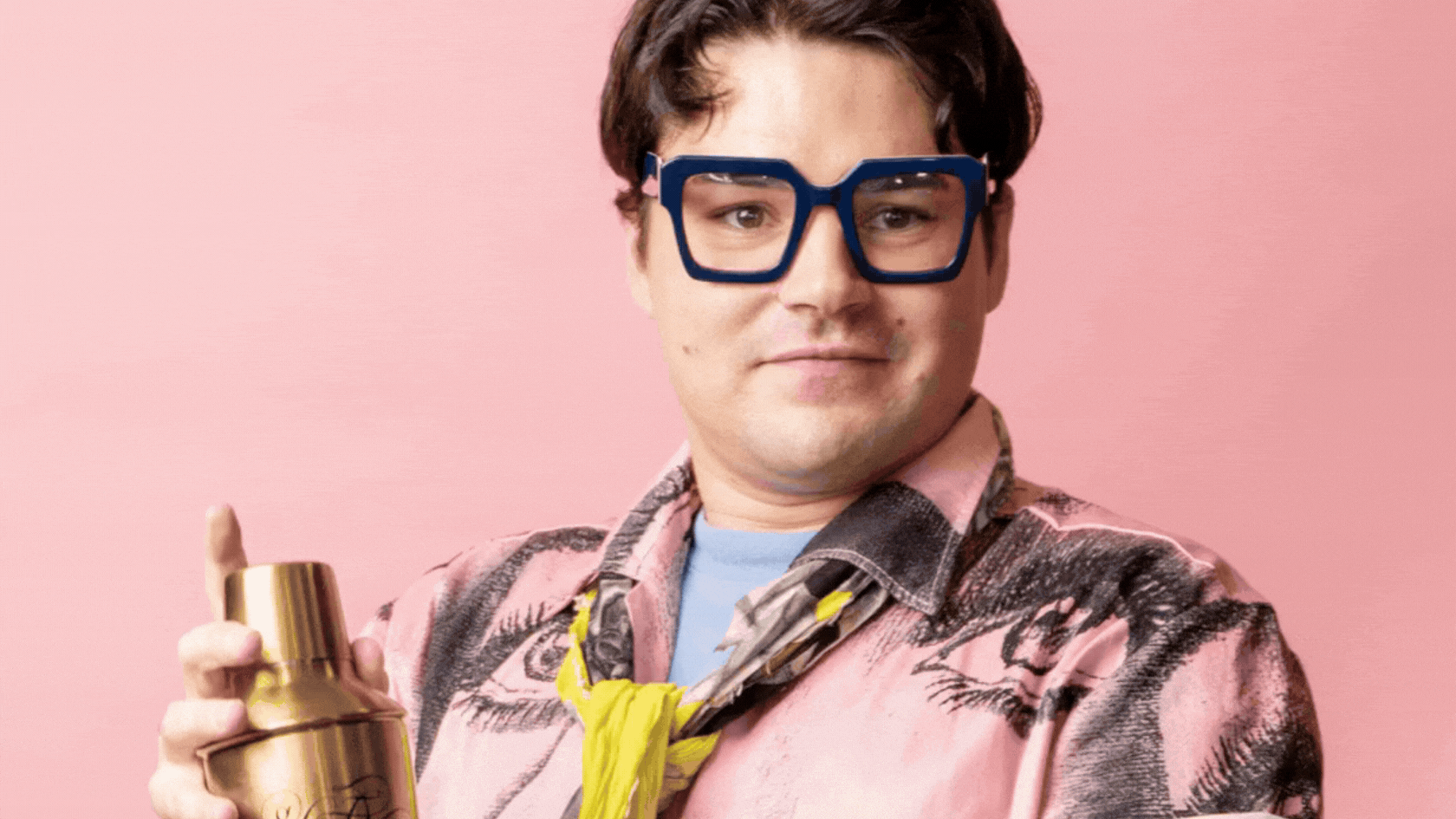 A gif of a bespectacled man wearing a pink shirt, yellow tie, and shaking a gold cocktail shaker